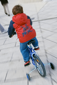A boy rides a bicycle on a paved footpath. Training wheels are attached to the bike.