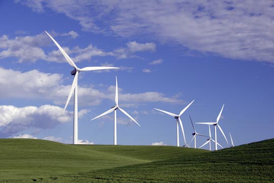 A photo of a green, slightly hilly landscape with a handful of wind turbines in the background.